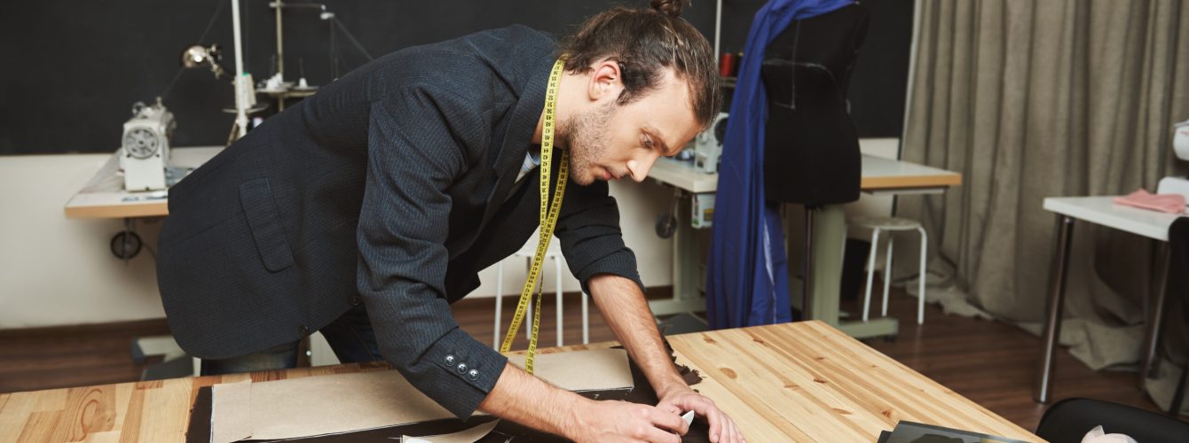Portrait of serious young good-looking hispanic fashion designer in black suit cutting out parts of jacket for winter collection with concentrated face expression